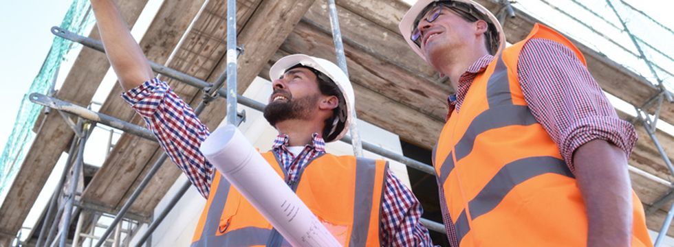 Zwei Handwerker mit Schutzhelm und Warnweste besprechen am Gerüst einen Bauplan