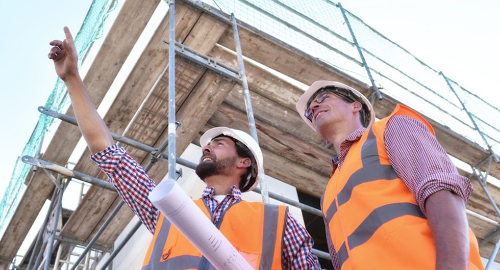Zwei Handwerker mit Schutzhelm und Warnweste besprechen am Gerüst einen Bauplan