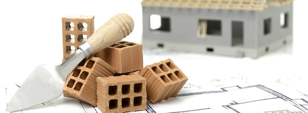 Ziegelsteine und eine Maurerkelle auf einem Bauplan vor einem Rohbaumodell